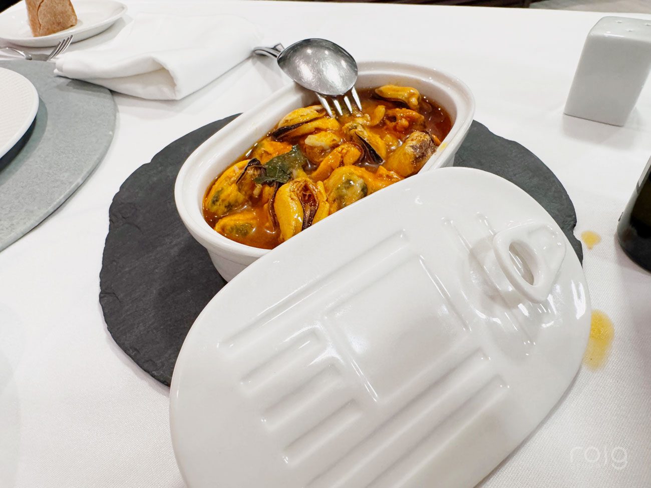 Cazuela de mariscos servida en recipiente cerámico blanco con tapa sobre plato de pizarra en restaurante El Mosquito
