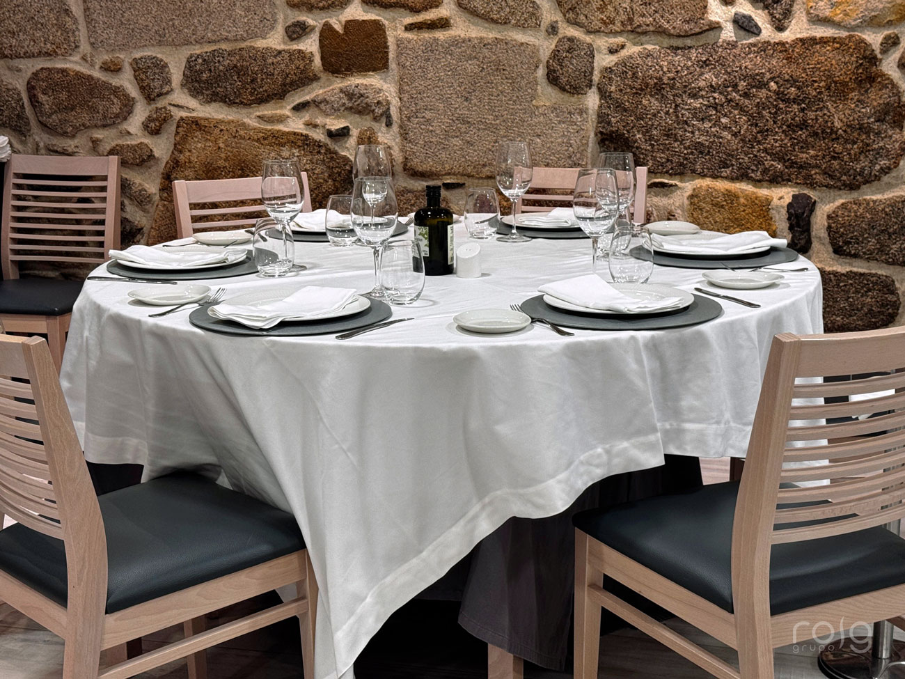 Mesa redonda con mantelería blanca, vajilla y cristalería completa frente a pared de piedra en comedor de El Mosquito